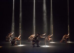 Six dancers on a black stage, crouched down, each holding a staff which is dramatically spotlight from above 