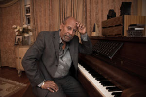 A smartly dressed older black gentleman at a piano in a domestic setting, turned away from the keys to look at the camera, left arm casually leaning on the piano with his hand on his head. It's a relaxed pose. He looks serious but friendly, perhaps a little quizzical (perhaps because he wants to play rather than pose for photos!)