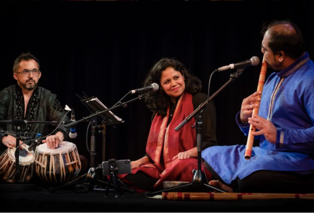 Shephali Frost, a woman of south Asian heritage sits on the floor in front of a microphone, with a black background. She is sitting in between and smiling at two male musicians, one with his hands rested on indian drums and another playing a large wooden flute.