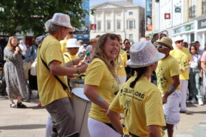 A photo of Emma from BrazilArte. A white woman with long blond hair who is smiling, dressed in a yellow t-shirt and white trousers, surrounded by others dressed in yellow t-shirts and white sparkly hats. Emma is in the middle of a pedestrianized high street, laughing and playing music as part of a street performance. 