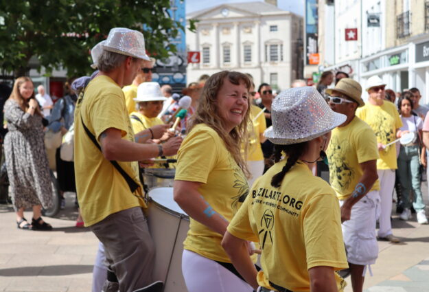 A photo of Emma from BrazilArte. A white woman with long blond hair who is smiling, dressed in a yellow t-shirt and white trousers, surrounded by others dressed in yellow t-shirts and white sparkly hats. Emma is in the middle of a pedestrianized high street, laughing and playing music as part of a street performance.