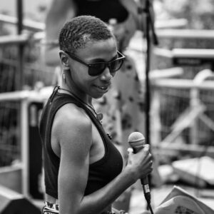 A black and white photo of TriggerBliss: a black woman with short hair and sunglasses, in a dark halter top. She is holding a microphone, her body facing away from the camera (like she is about to go on stage) but head is turned, and she is glancing back towards us with a smile. 