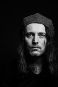 A black and white photo of Ian: a white man with long dark hair and a thin goatee beard / stubble, wearing a dark tweed beret. Ian is looking into the camera with a calm but serious expression on his face.