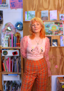 A picture of Lu in their studio: a white woman with yellow and orange haie, dressed in bright clothing, a pink top and tartan trousers, smiling at the camera.
