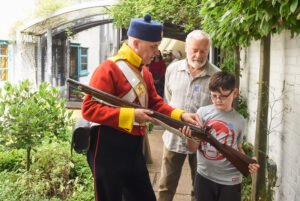 A photo of a white man in a victorian soldier's costume, showing a gun to a male child aged around 10, and an older man behind.
