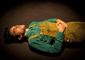 A photo of Vijay taken during a live performance of their show, Sometimes I Leave. Vijay is a brown person with blue hair, wearing a leafy green shirt and yellow washed dungarees. They are laying on a black stage floor with their eyes closed. A general lighting wash is covering the stage