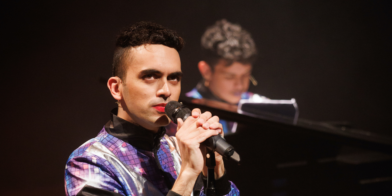 A photo of Vijay Patel. Vijay is a brown person holding a microphone and wearing a purple top, with a ypoung person, slightly blurred in the image, behind them playing piano