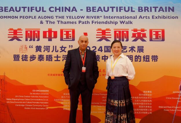 A picture of our CEO Indi Sandhu with Simone Xue, ECDP Trustee and founder of Colchester Chinese Culture Society. Indi is of south asian heritage dressed in a suit. Simone is of Chinese heritage, is wearing a white blouse with a black dress with a gold pattern. They are standing in front of a huge colourful poster with Chinese writing.