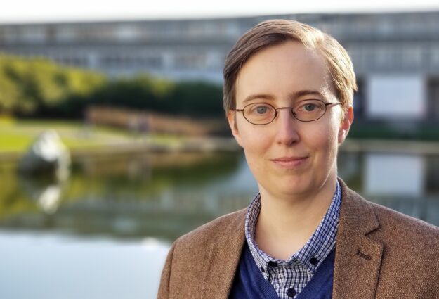 Small, white human with mousy brown short hair and glasses. In this photo I am wearing a brown tweed jacket, and a blue jumper with a checked shirt collar. Standing in front of a body of water and brutalist building where Battlestar Galactica was filmed, which unless you are as geeky as me you will only know if you read this alt-text!