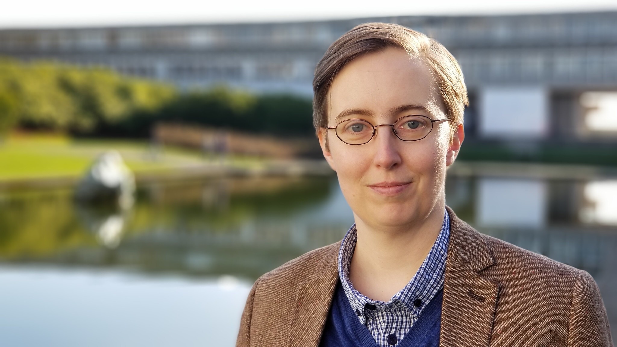 Small, white human with mousy brown short hair and glasses. In this photo I am wearing a brown tweed jacket, and a blue jumper with a checked shirt collar. Standing in front of a body of water and brutalist building where Battlestar Galactica was filmed, which unless you are as geeky as me you will only know if you read this alt-text!