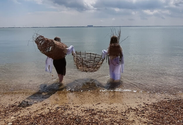 Two people on a beach, walking away from the camera into the sea, carrying wicker fishing baskets.