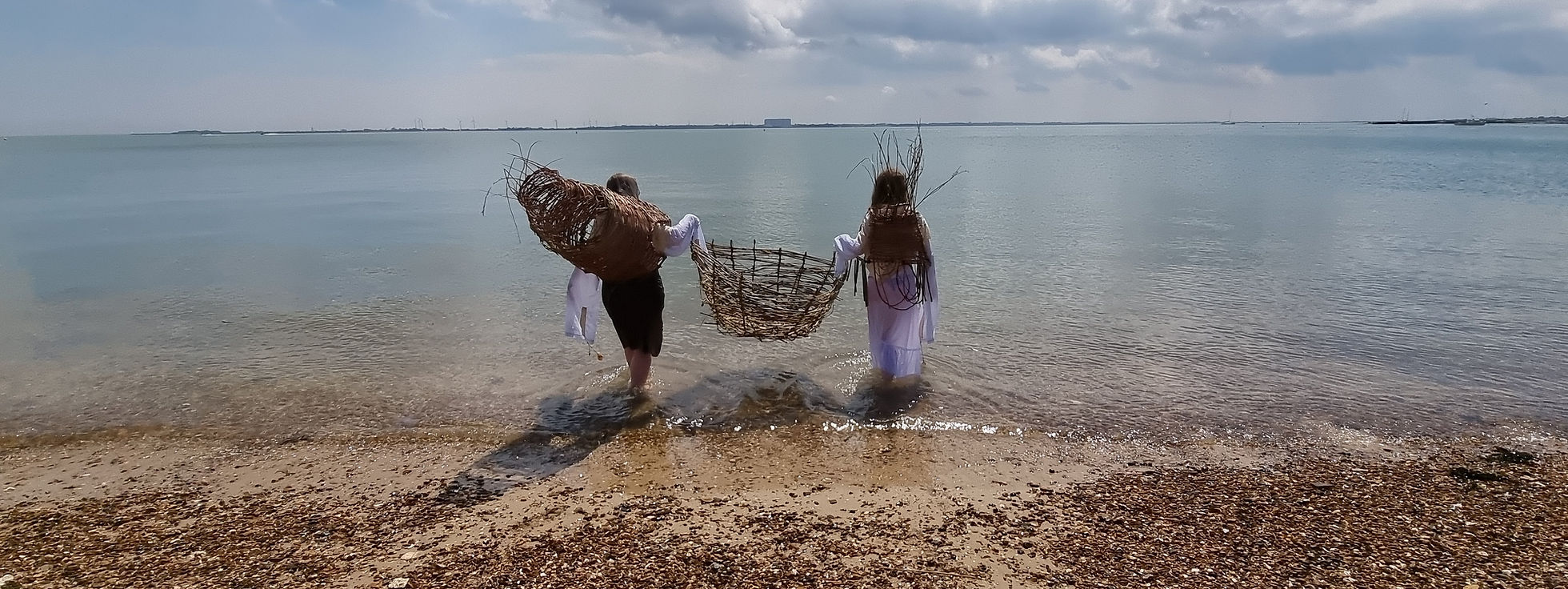 Two people on a beach, walking away from the camera into the sea, carrying wicker fishing baskets.